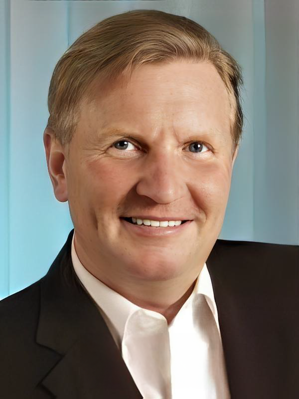 Mann mit blonden Haaren lächelt in die Kamera, trägt eine schwarze Jacke und ein weißes Hemd auf blauem Hintergrund.