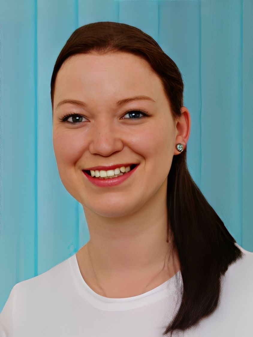 Frau mit braunem Haar im Pferdeschwanz lächelt; hellblauer Hintergrund.