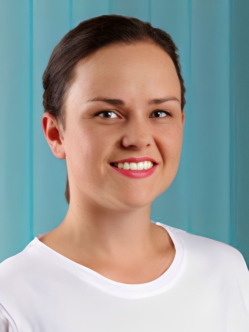 Frau mit dunklem Haar, lächelnd, trägt ein weißes Hemd vor hellblauem Hintergrund.