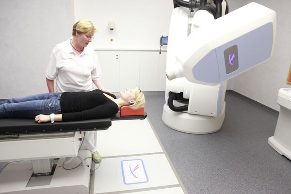 Frau erhält Strahlentherapie; medizinisches Personal richtet den Kopf der Patientin. Raum mit Roboterarm.