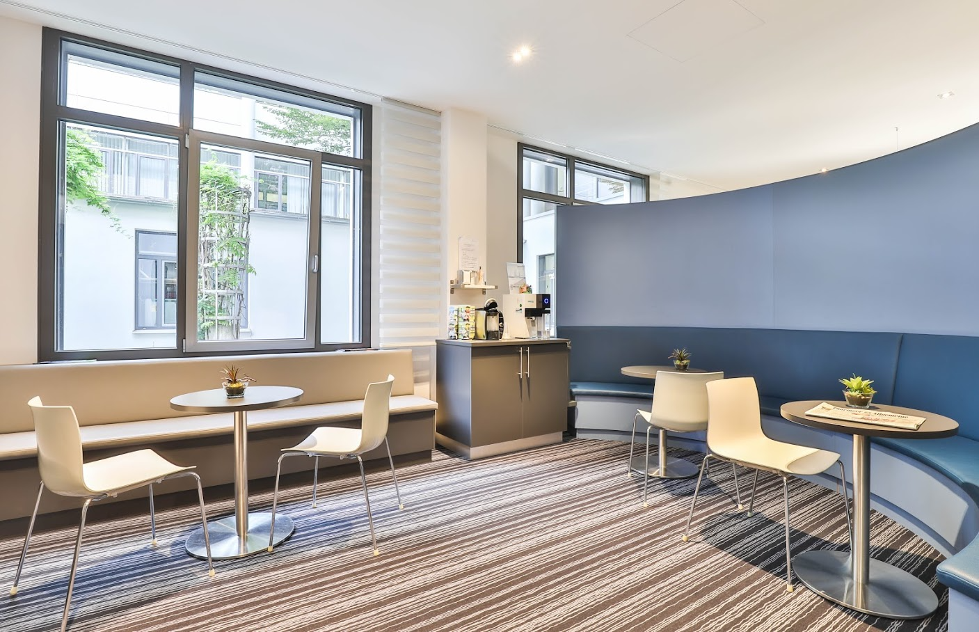 Loungebereich mit Tischen, Stühlen, eingebauten Sitzgelegenheiten, Kaffeestation und Fenstern mit Blick auf einen Innenhof.