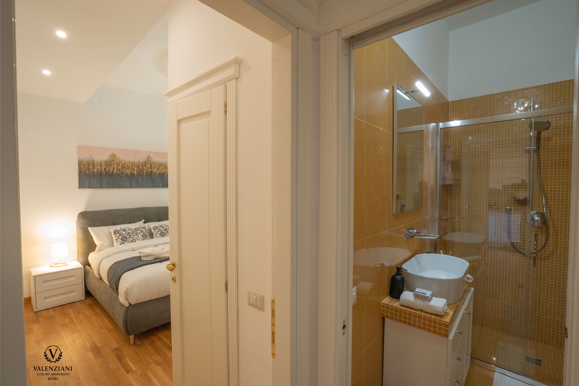 Bedroom and bathroom view. White doors, grey bed, yellow-tiled shower, white sink.