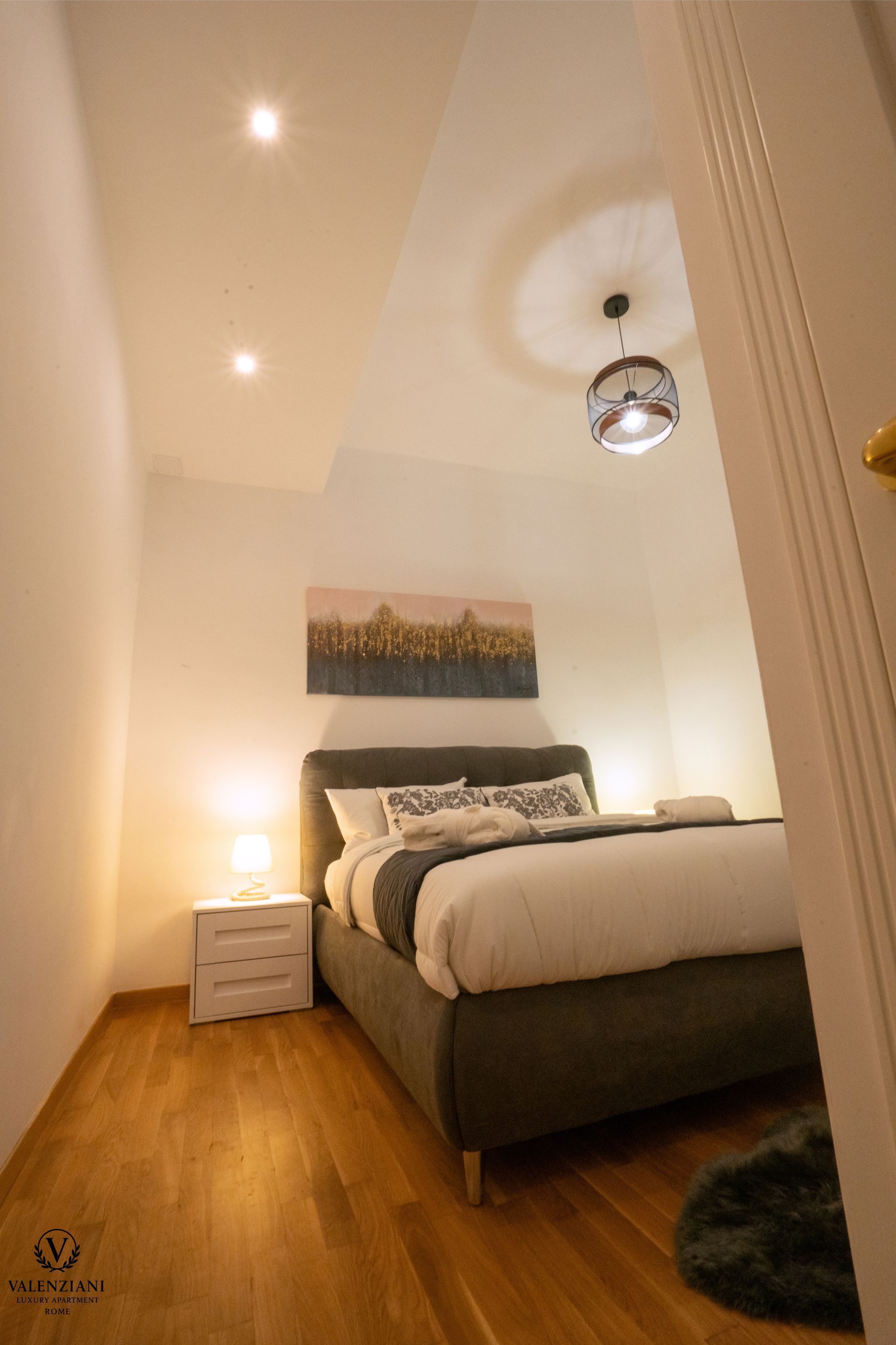 Cozy bedroom with a bed, nightstand, artwork, and overhead lighting; wood flooring and white walls.