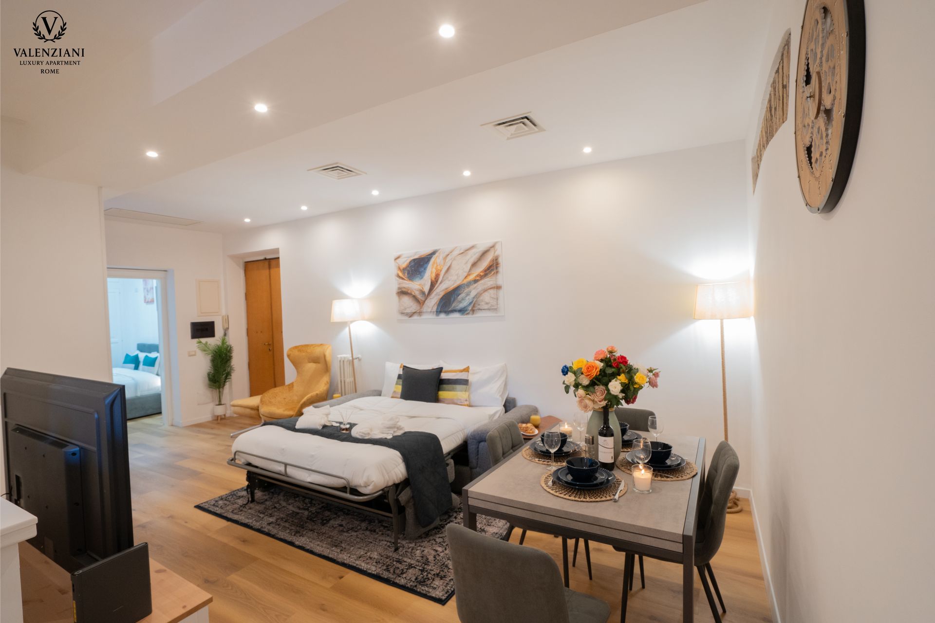 A well-lit studio apartment with a bed, dining table, and TV. White walls, wooden floor, and a vibrant flower arrangement.