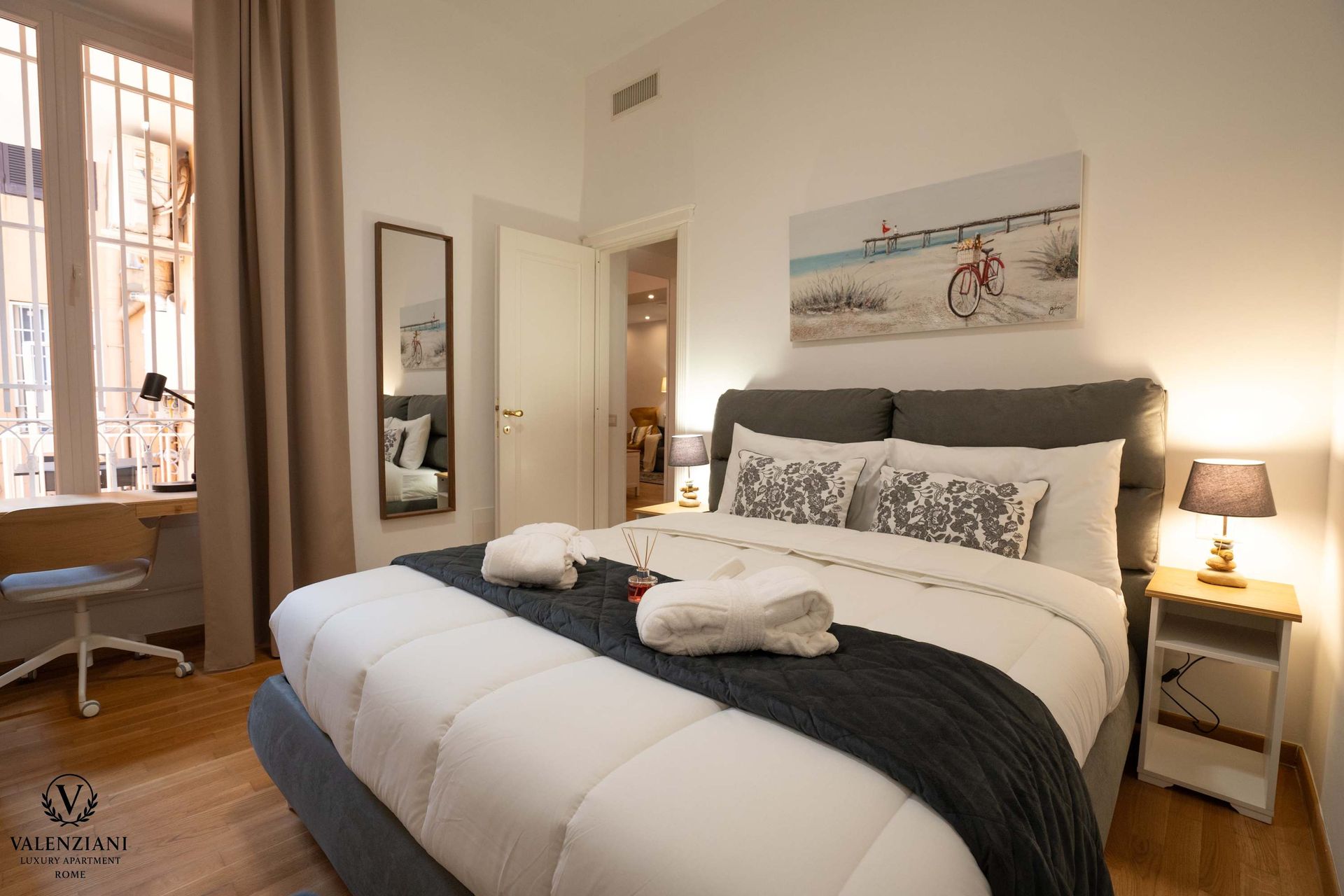 Bedroom with a bed, mirror, and window. Bed has white linens, gray throw and pillows. Small desk by window.