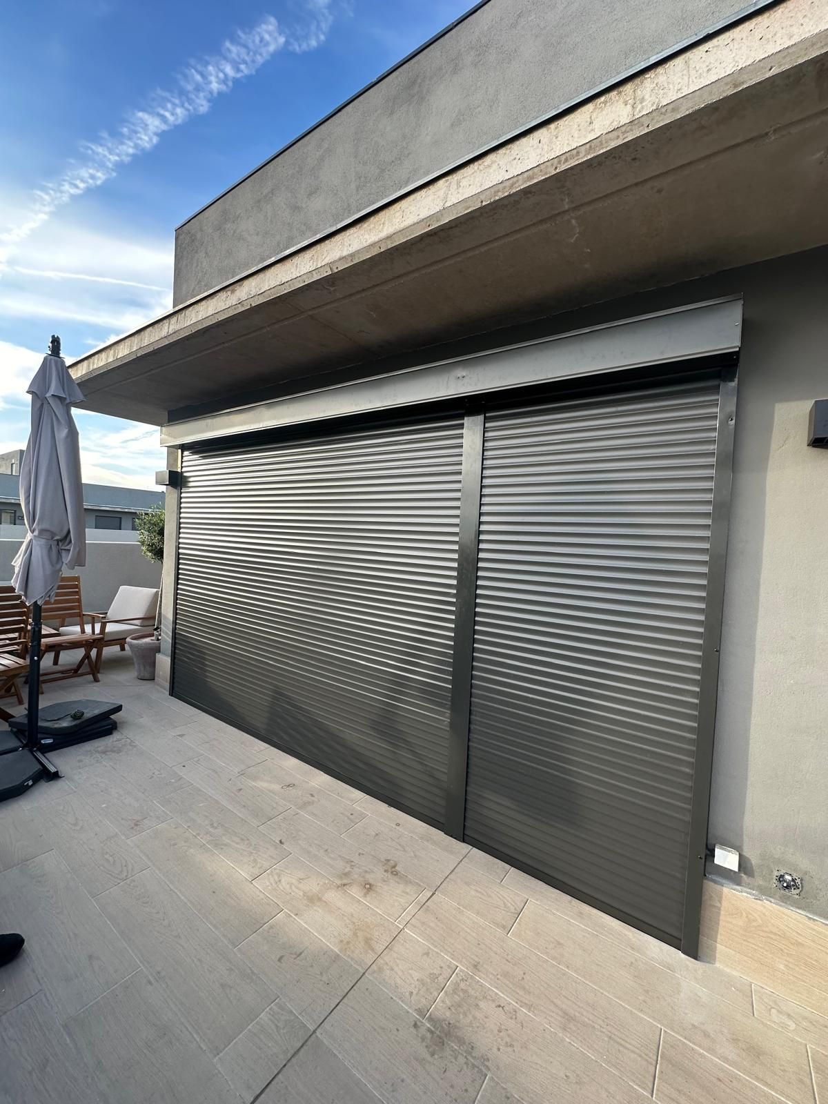 Vista exterior de un patio con persianas grises cerradas. Se ven el patio gris, la sombrilla y los muebles.