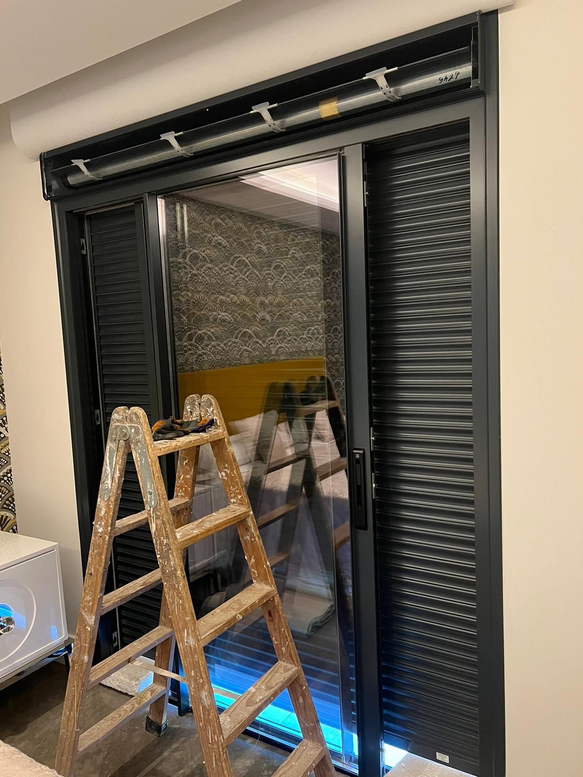 Una puerta corrediza de vidrio con marco oscuro con una contraventana cerrada, una contraventana abierta y una escalera de madera en el interior.