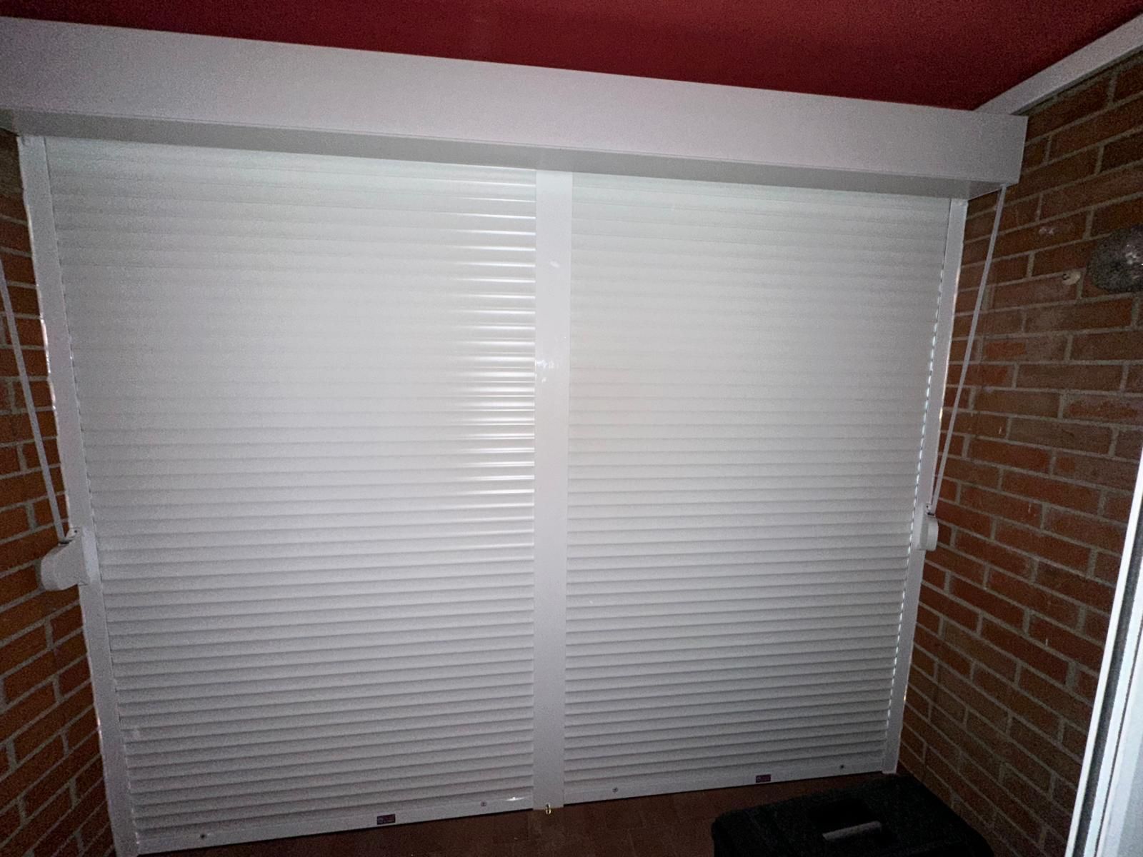 Persianas blancas cubren una ventana en una pared de ladrillo. Se ve un techo rojo por encima de las persianas.