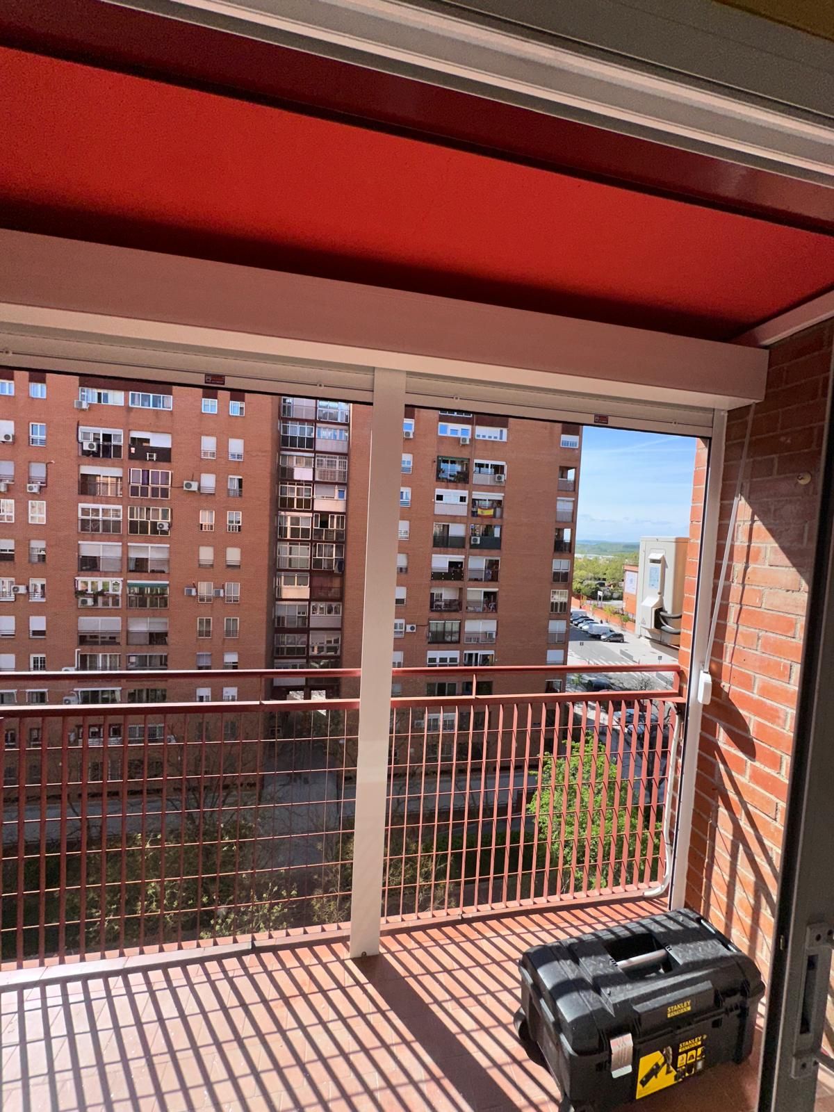 Balcón con toldo rojo, barandilla metálica, vista a edificios y caja de herramientas.