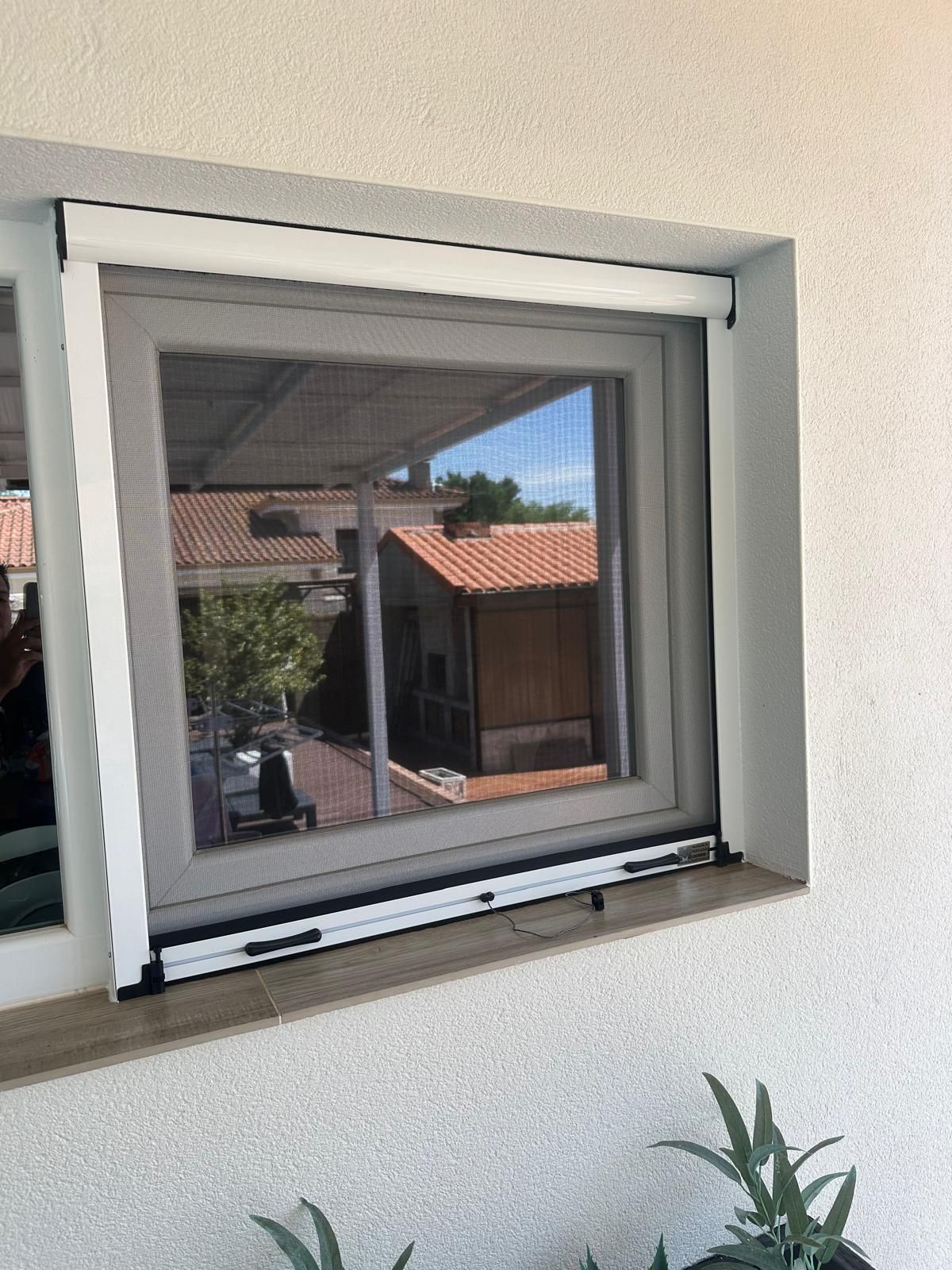 Una ventana con pantalla retráctil, ubicada en una pared blanca; la vista incluye un techo con tejas, entorno al aire libre.