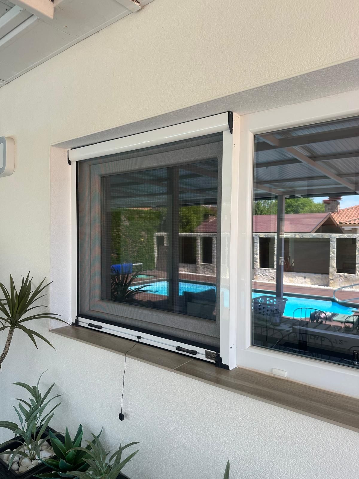 Ventana con marco blanco y pantalla retráctil; vista exterior de un área de piscina.