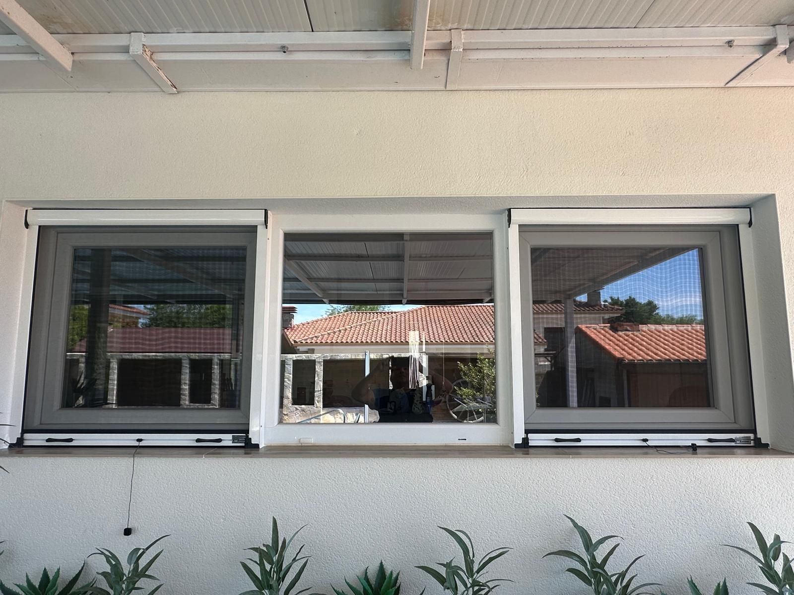 Tres ventanas con marcos blancos y mosquiteros retráctiles, situadas en una pared exterior blanca, sobre plantas verdes.