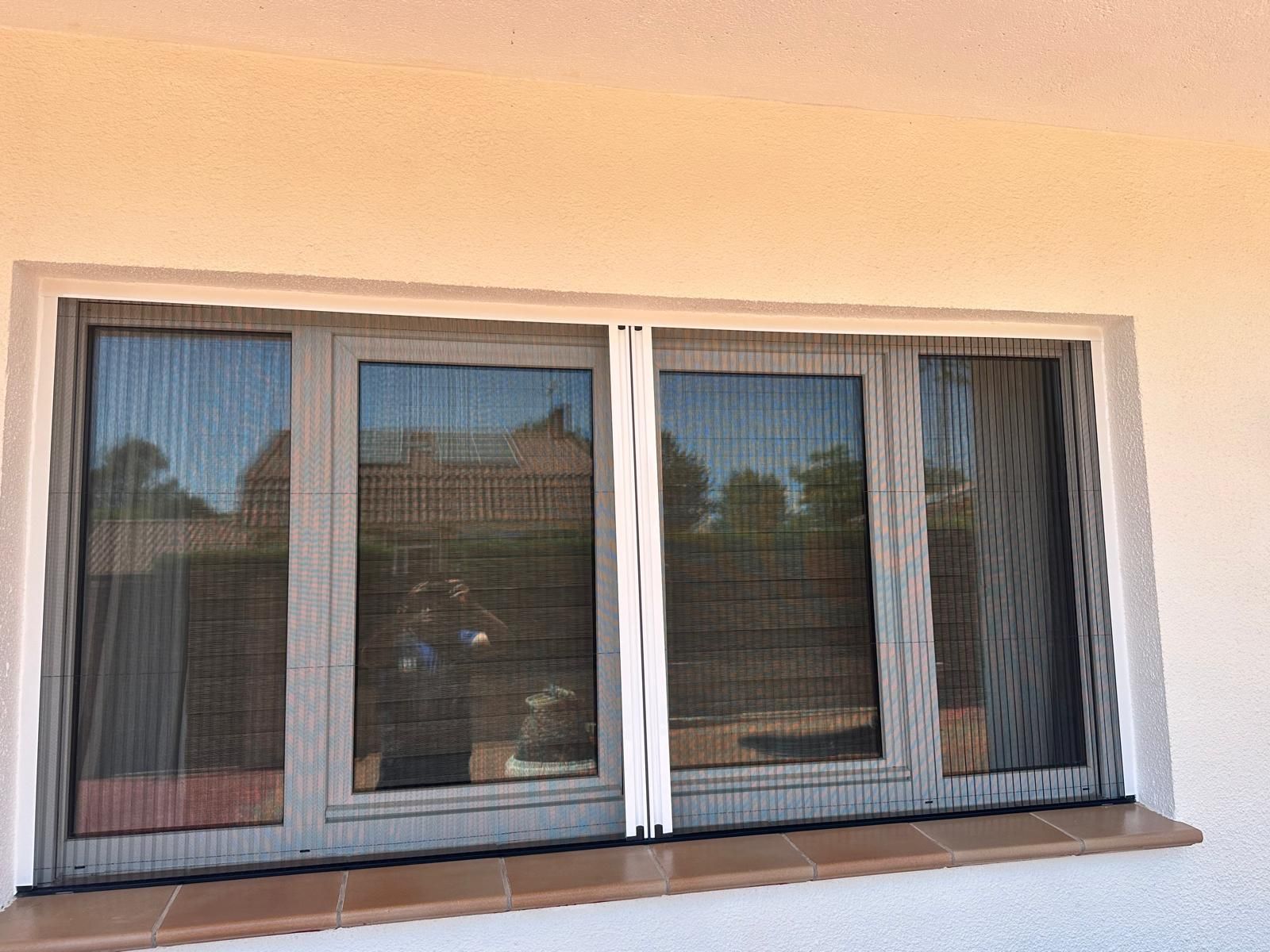 Una ventana con una malla negra, colocada en un marco blanco, contra una pared de color claro.