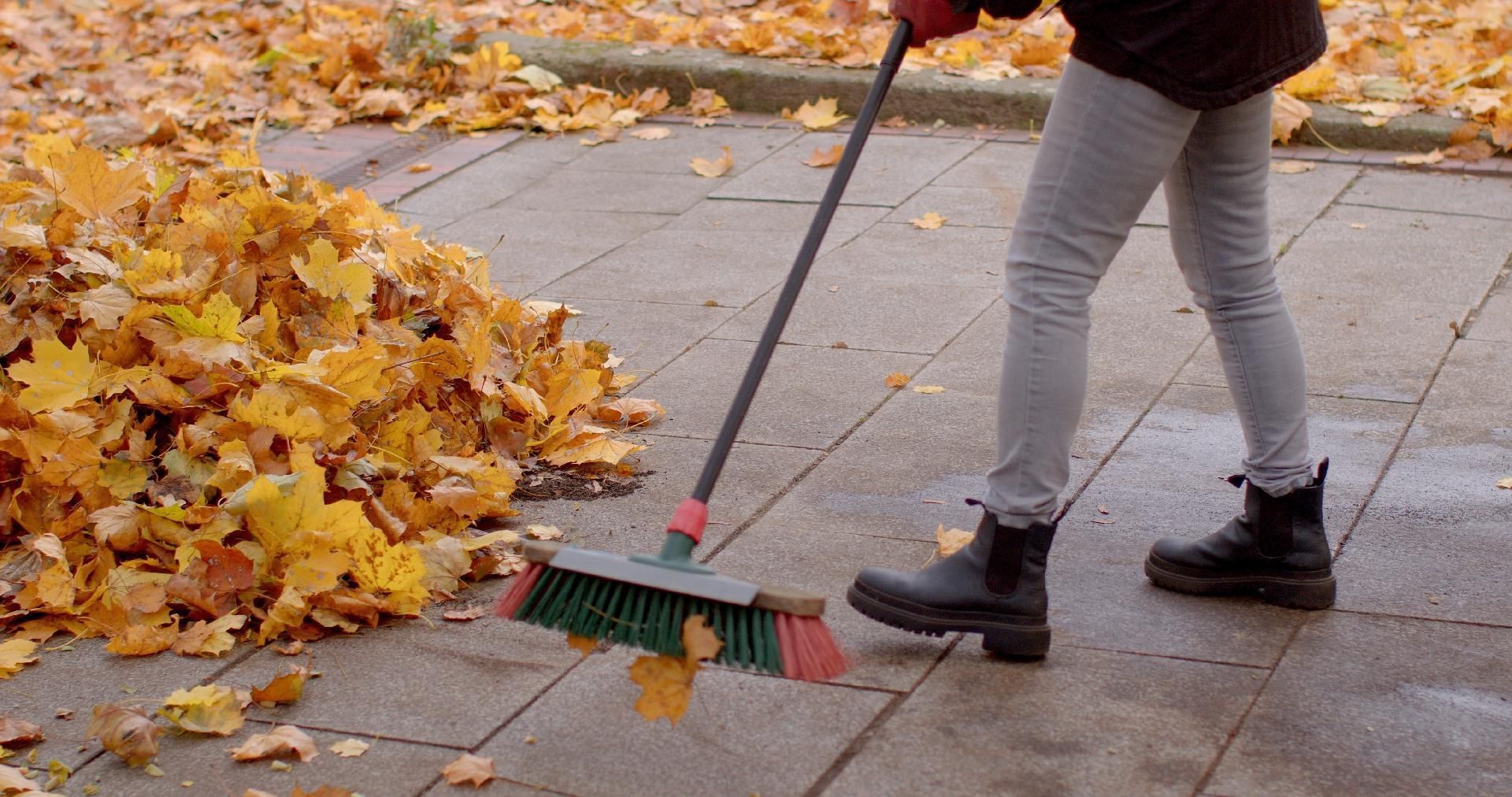 Person fegt Herbstlaub von Gehweg mit Besen