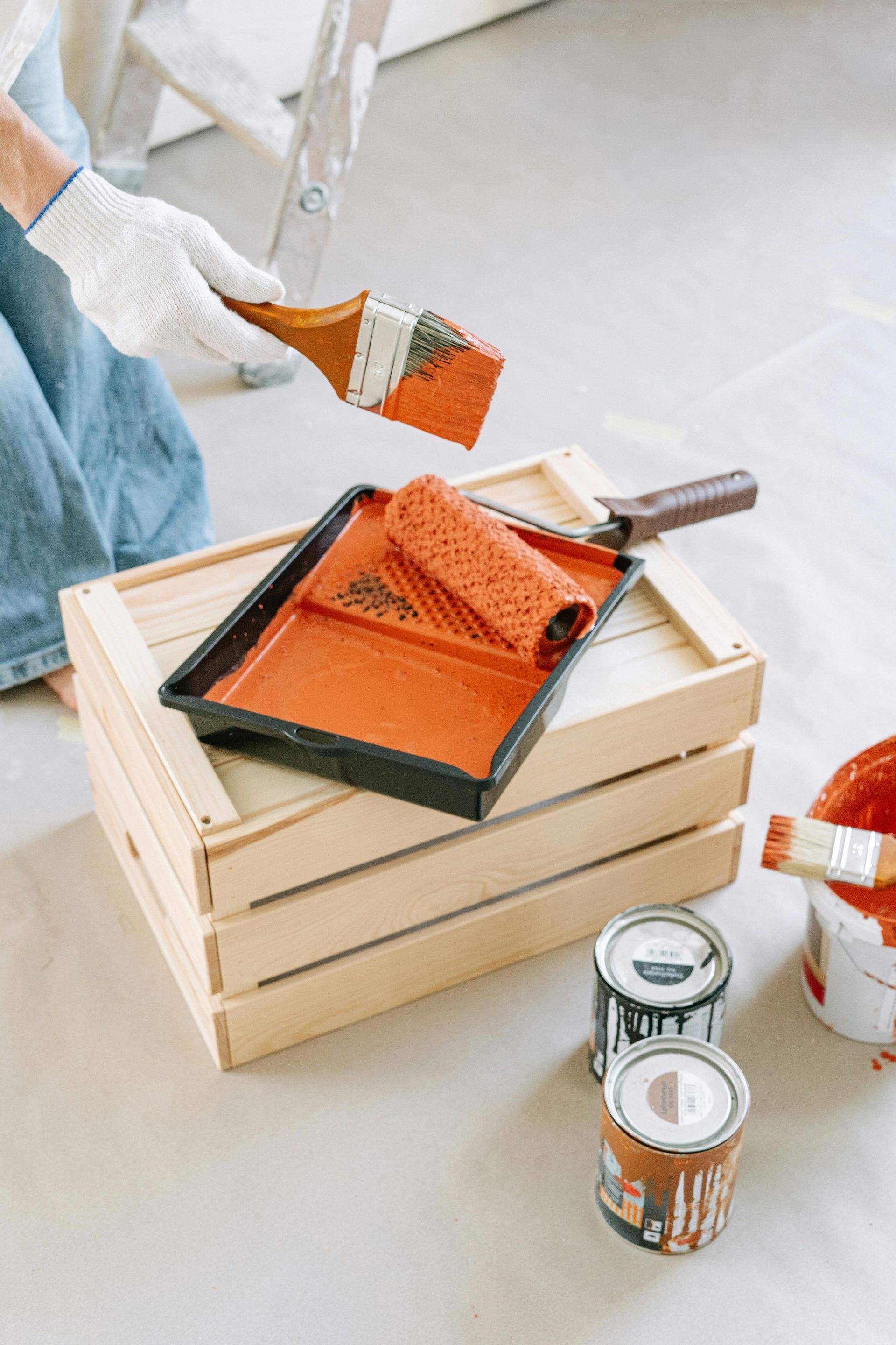 Bandeja para rodillo de pintura con pintura naranja sobre cajas de madera apiladas, con latas de pintura cerca.