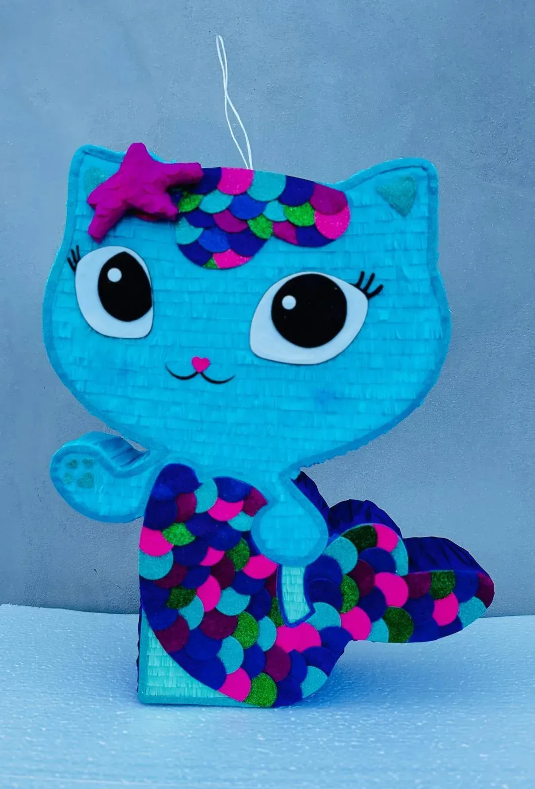 Adorno de gato azul de dibujos animados con detalles de lunares, ojos grandes y un lazo, colgando sobre un fondo azul.
