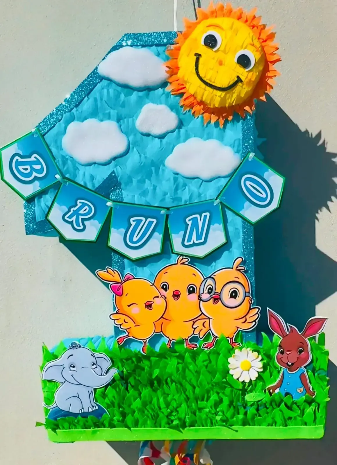 Manualidad colorida “BRUNO” con sol, nubes, hierba y animales de dibujos animados sobre un fondo azul.