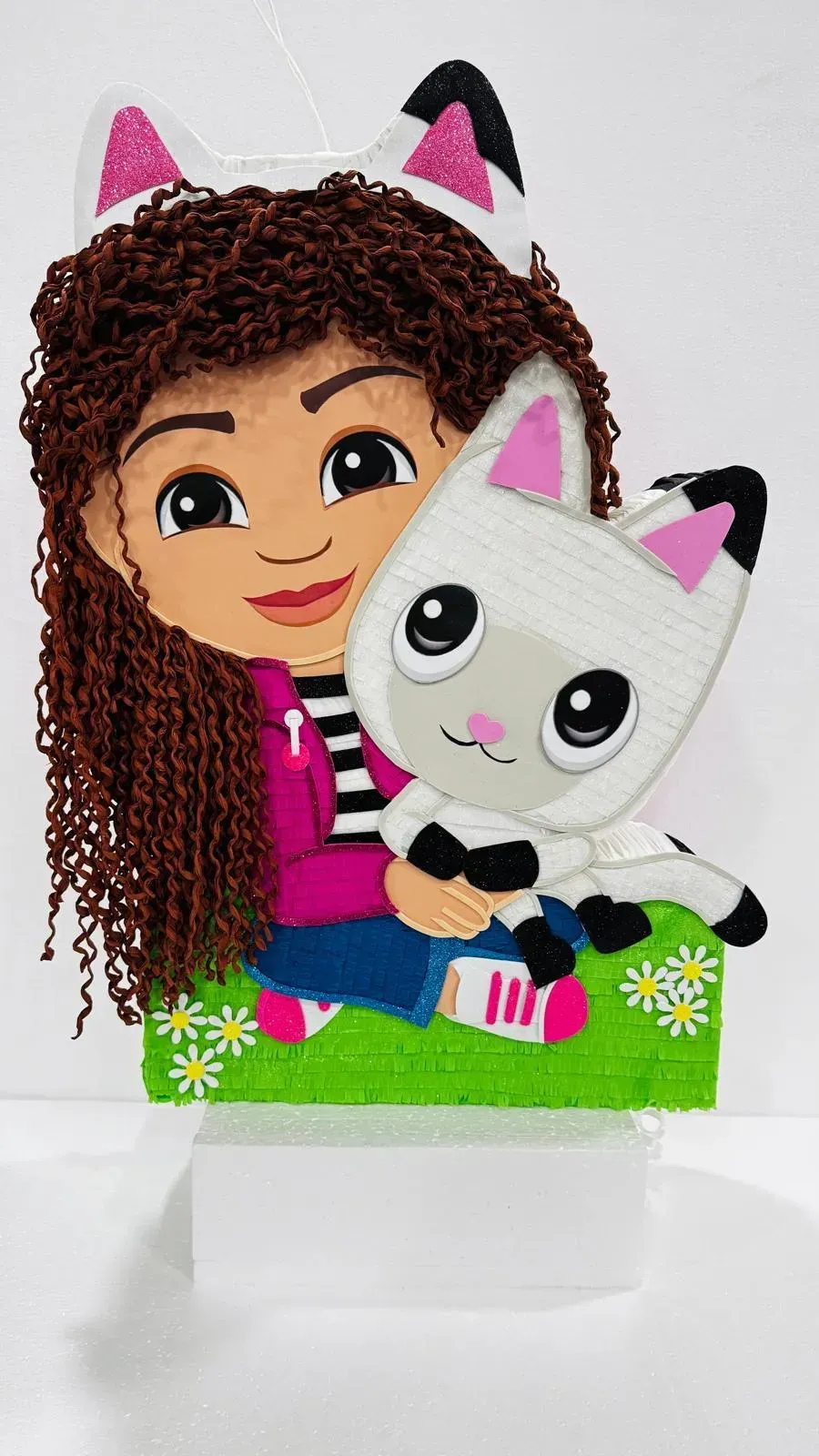 Niña de dibujos animados sosteniendo un peluche de gato blanco con orejas rosas, sentada sobre una base verde.
