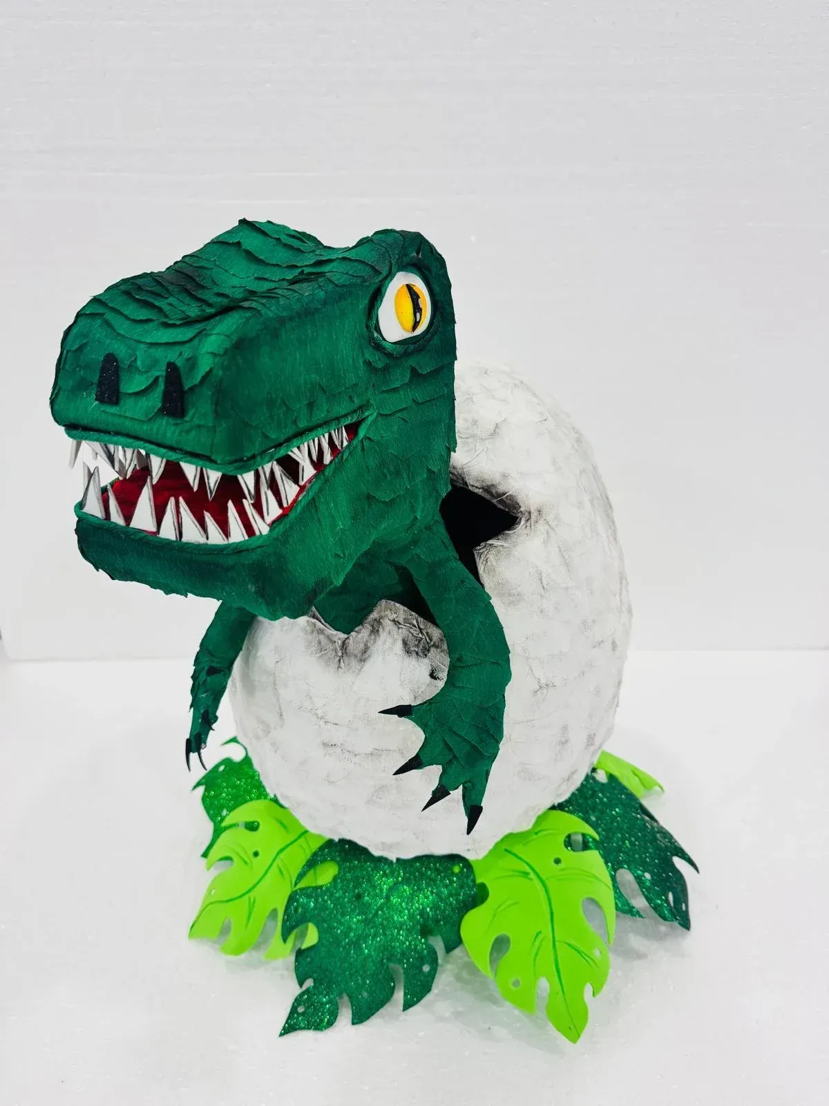 Dinosaurio de juguete verde en una cáscara de huevo blanca con patas de color verde brillante y ojo amarillo, sobre un fondo liso.