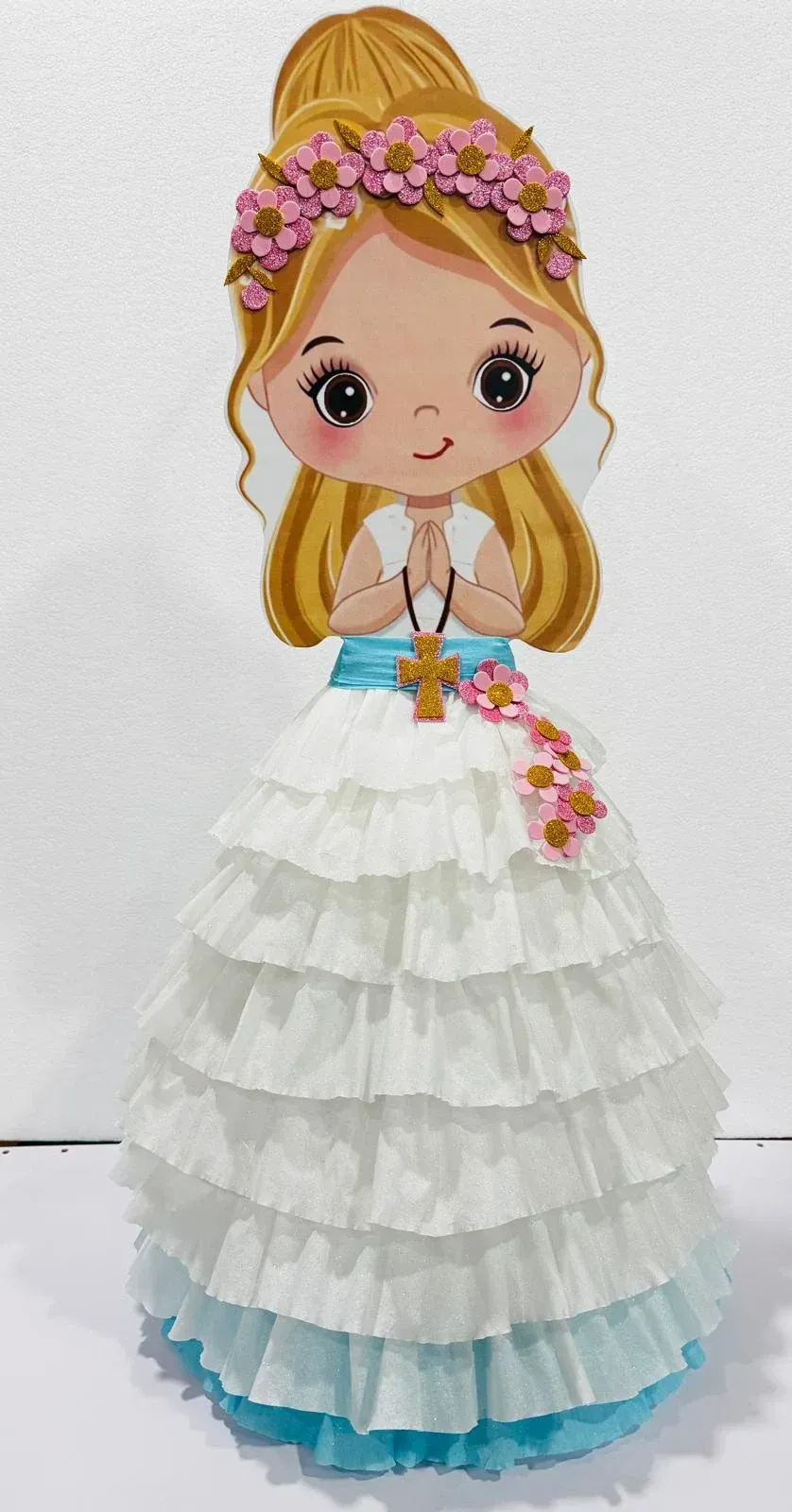 Muñeca ilustrada con un vestido blanco con volantes, flores rosas y dobladillo azul, y un tocado floral.