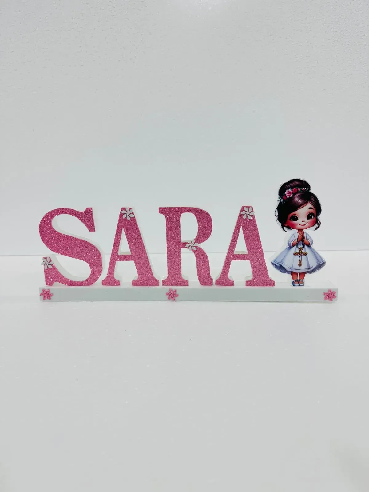 Diseño con el nombre “SARA” en purpurina rosa, con una pequeña niña de dibujos animados y estrellas sobre fondo blanco.