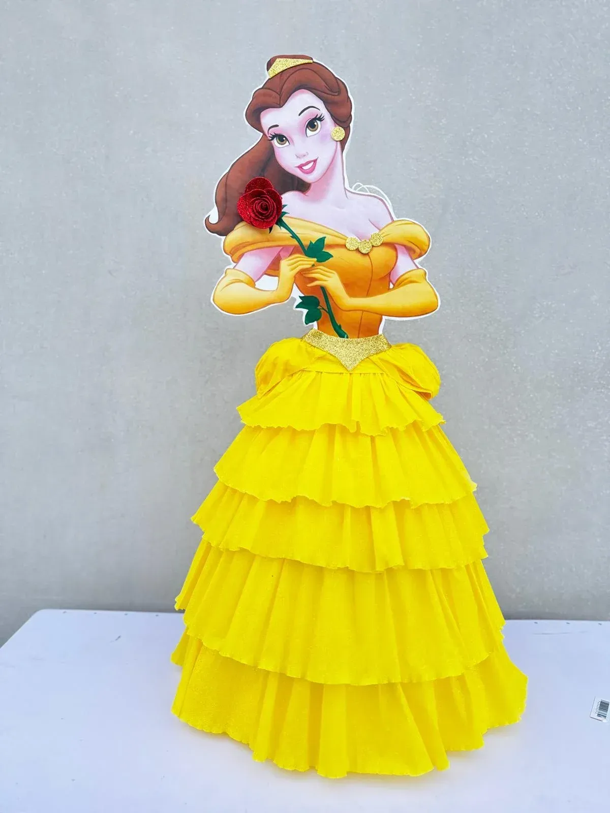Figura de Bella con un vestido amarillo brillante sosteniendo una rosa roja, de pie sobre un fondo liso.