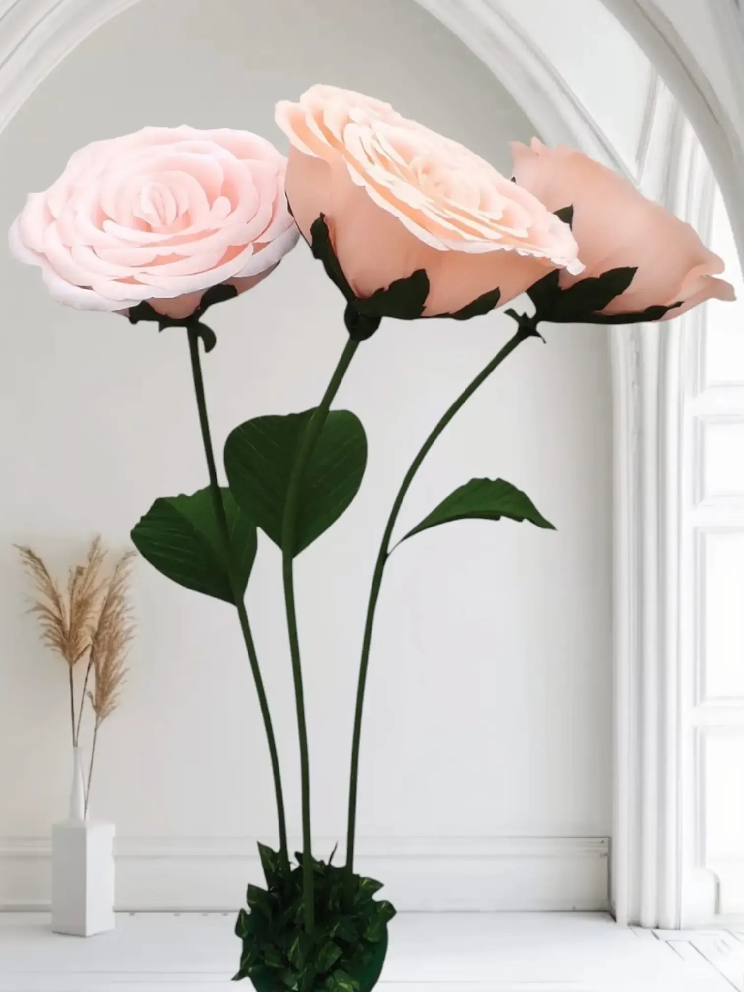 Tres rosas de color rosa pálido con tallos largos en un jarrón blanco junto a una ventana luminosa.