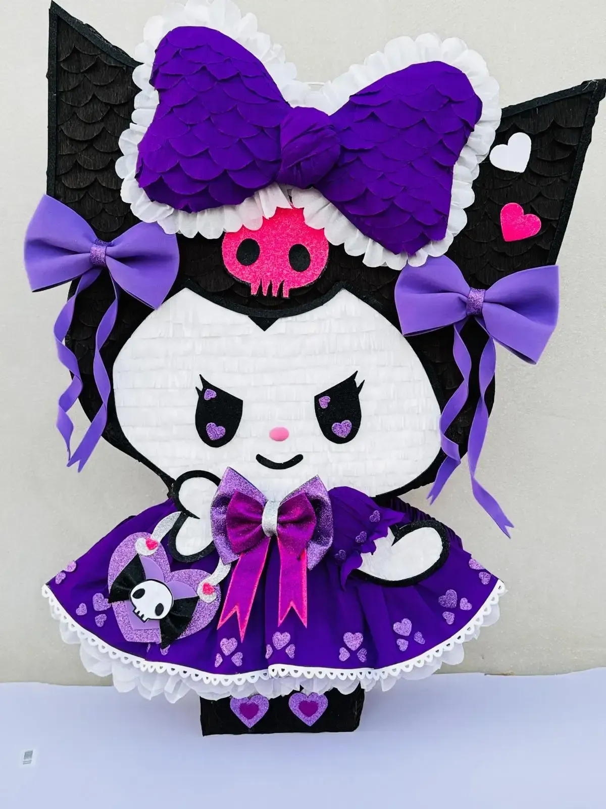 Figura de Kuromi de color morado y negro con lazos, detalles de calaveras y un vestido con volantes sobre un fondo blanco.