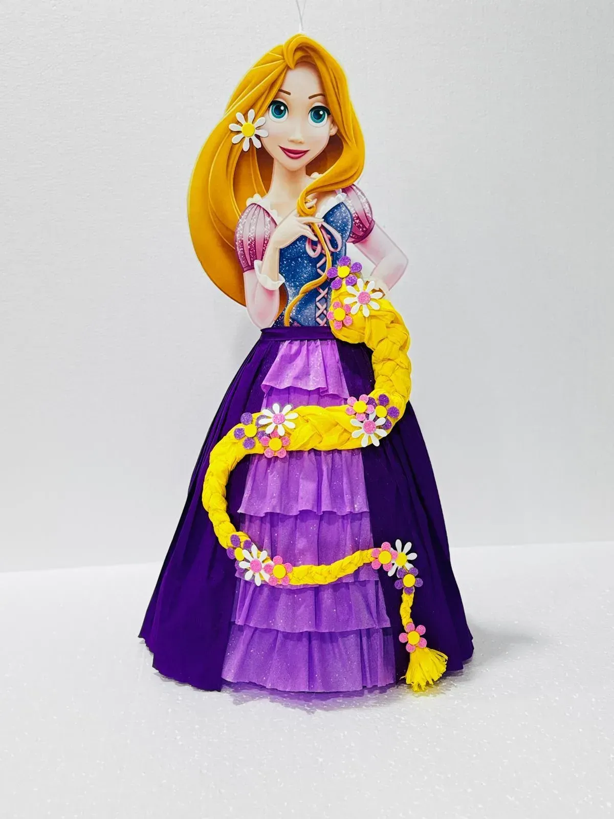 Una muñeca vestida de morado con cabello rubio largo y una guirnalda de flores amarillas sobre un fondo blanco liso.