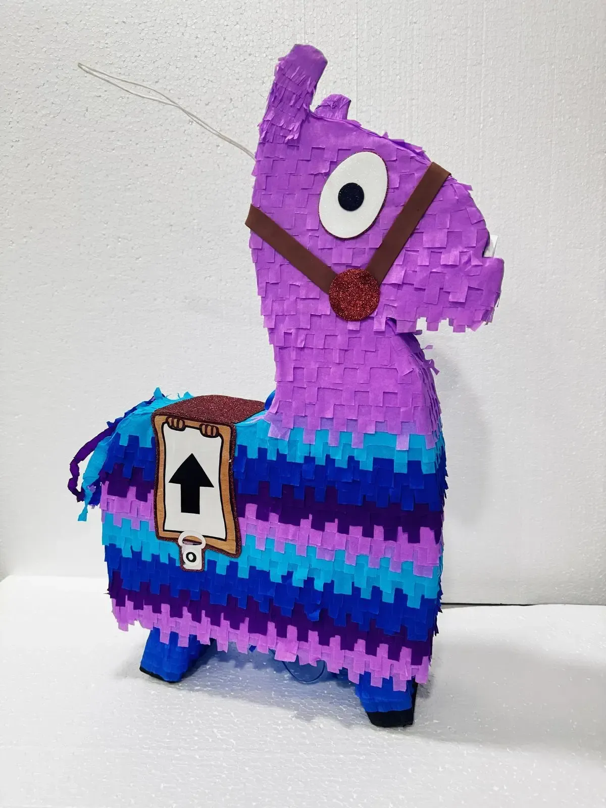 Piñata de llama morada con rayas azules, silla de montar marrón y una etiqueta numerada sobre un fondo blanco.