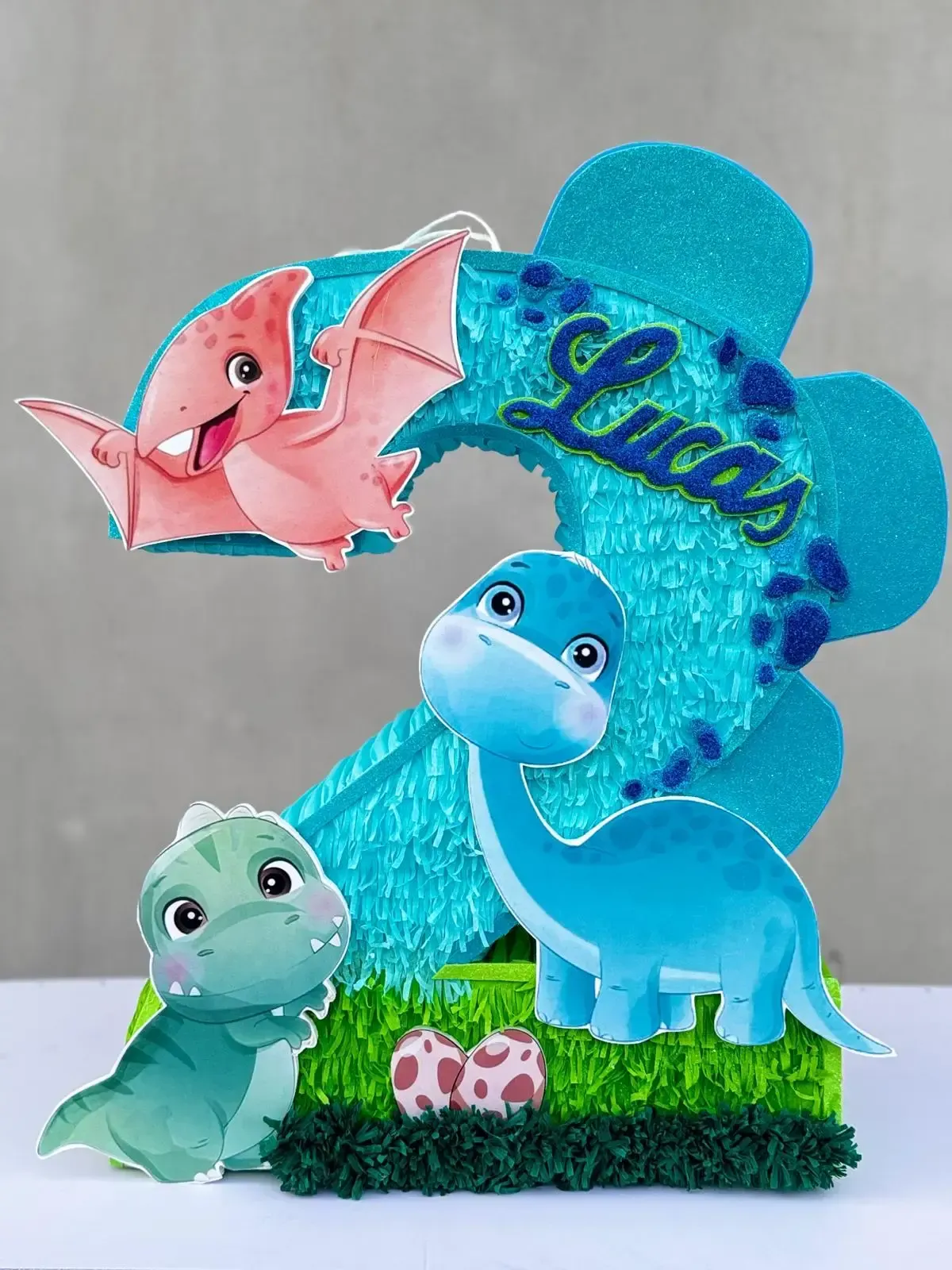 Tres figuras de dinosaurios de dibujos animados en tonos pastel sobre el césped, con un letrero azul brillante que dice 