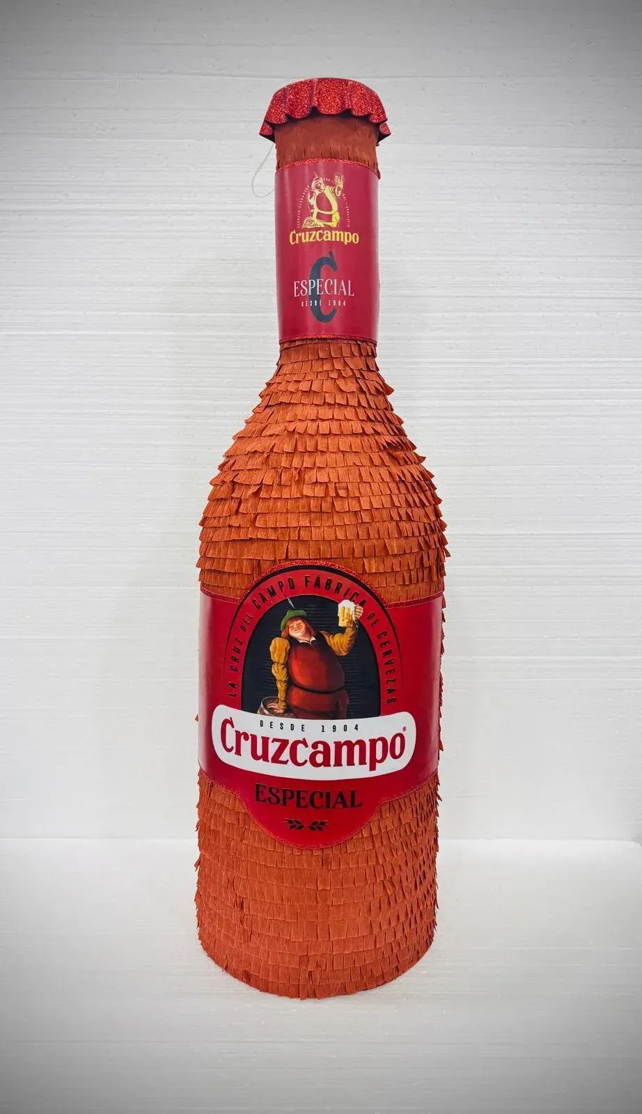 Botella de cerveza Cruzcampo envuelta en material texturizado naranja sobre un fondo blanco.