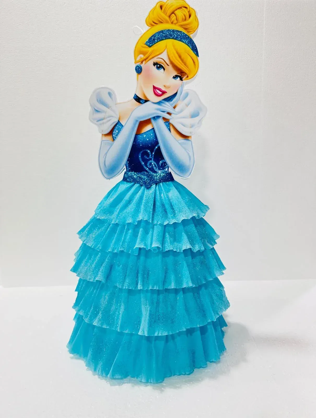 Muñeca Cenicienta azul con un vestido brillante y un moño rubio, de pie sobre un fondo blanco.