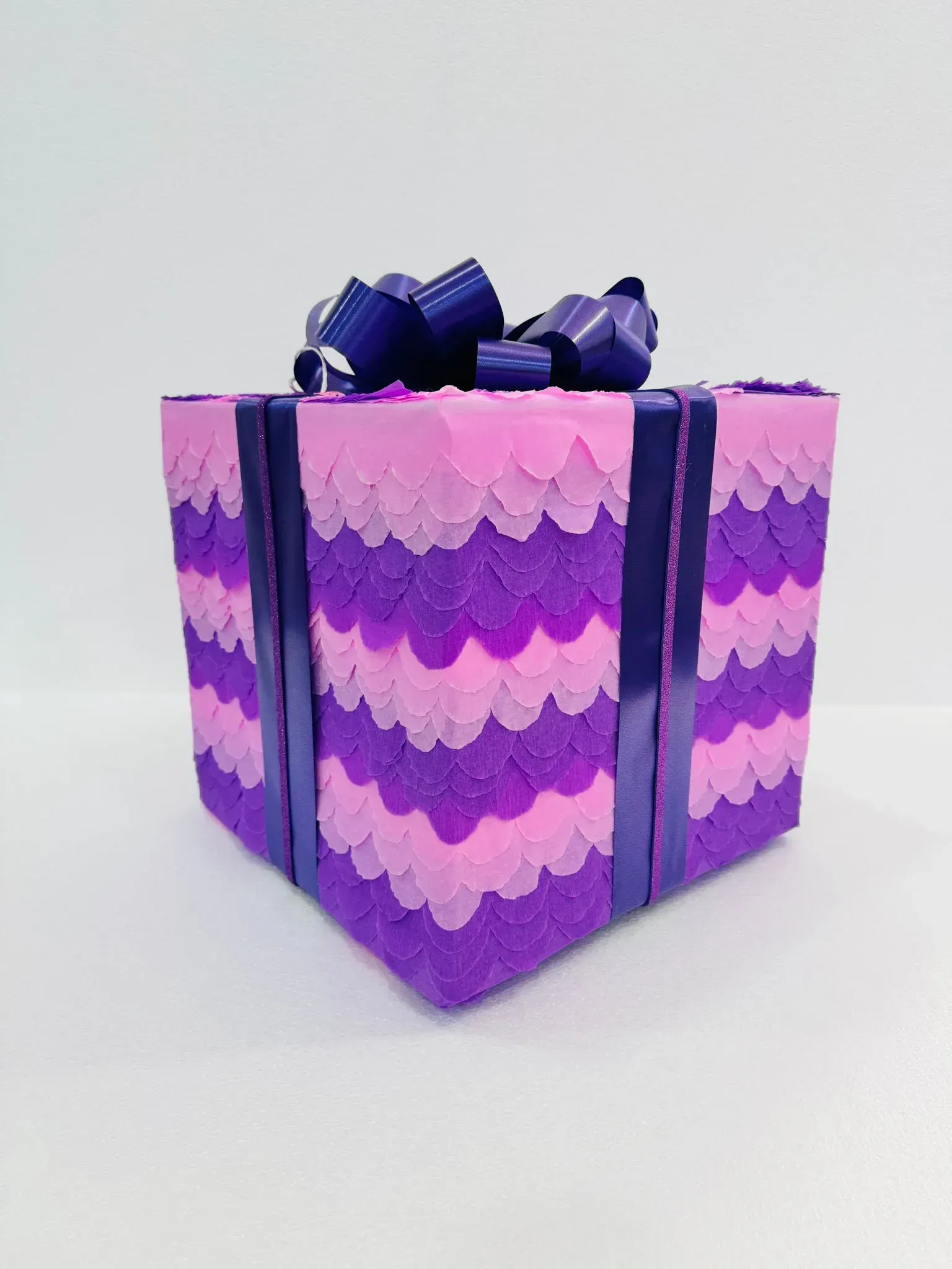 Caja de regalo morada con borde festoneado, cinta oscura y lazo sobre fondo blanco.