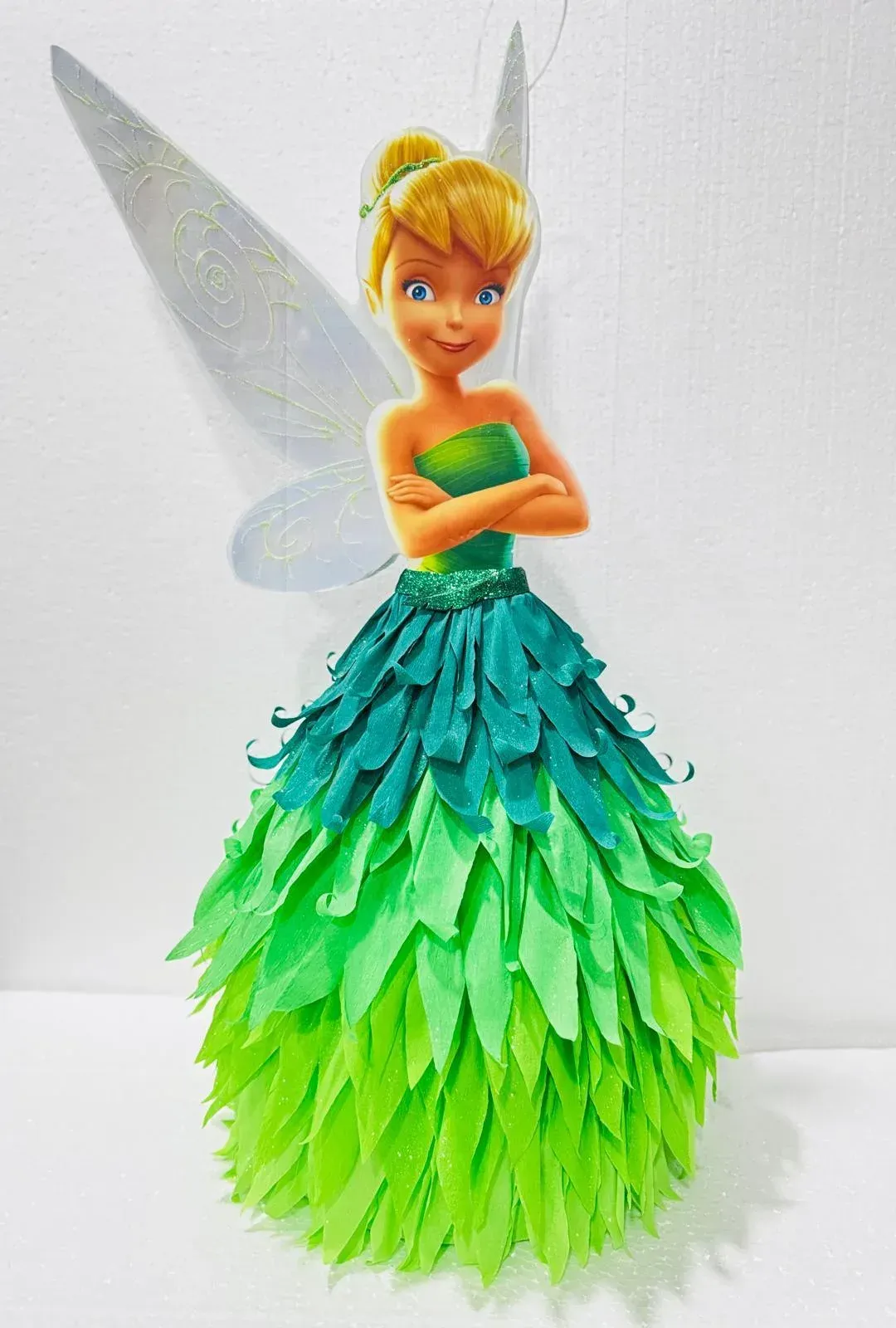 Figura de Campanilla con un vestido de plumas verdes y alas, de pie sobre un fondo blanco.
