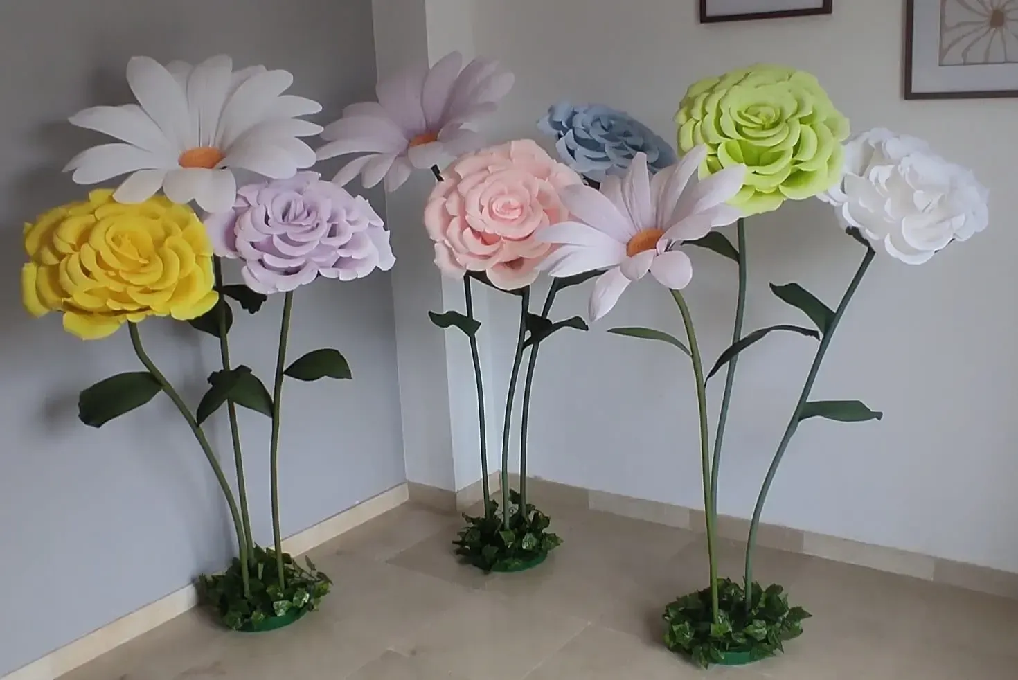 Flores de papel decorativas de gran tamaño en amarillo, morado, rosa, azul, verde y blanco sobre soportes en interiores.