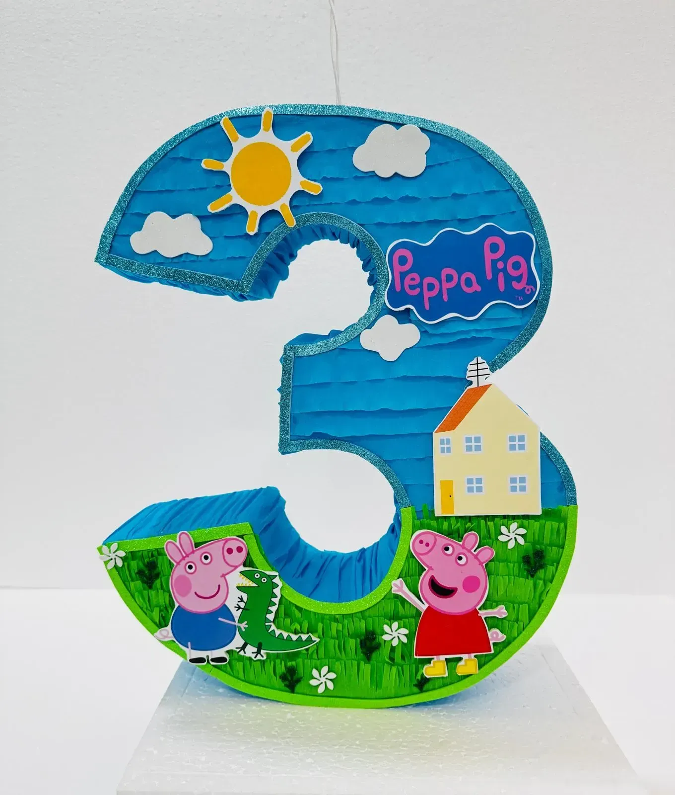 Decoración con el número 3 inspirada en Peppa Pig, con cielo soleado, césped, casas y personajes de dibujos animados.
