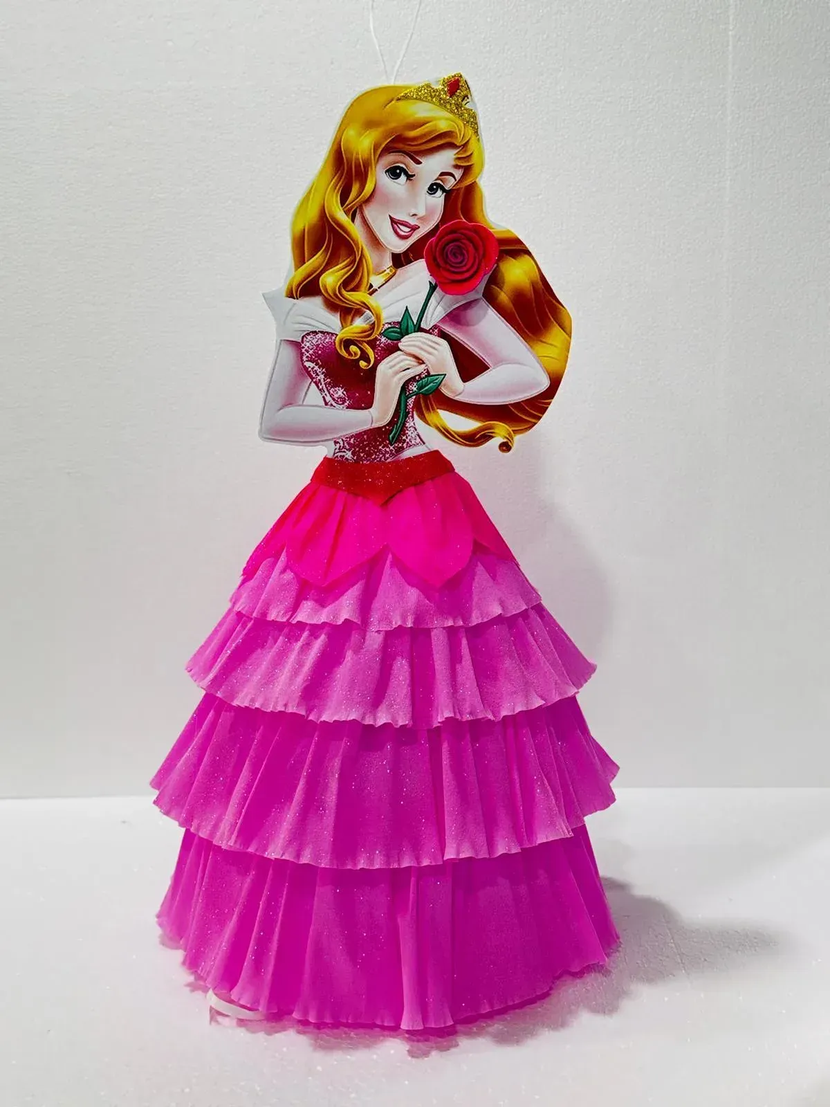 Muñeca princesa rosa con un vestido de capas, sosteniendo una rosa roja y de pie sobre un fondo blanco.