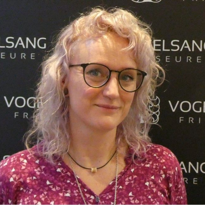 Eine Frau mit lila Haaren trägt ein schwarzes Hemd und lächelt in die Kamera.