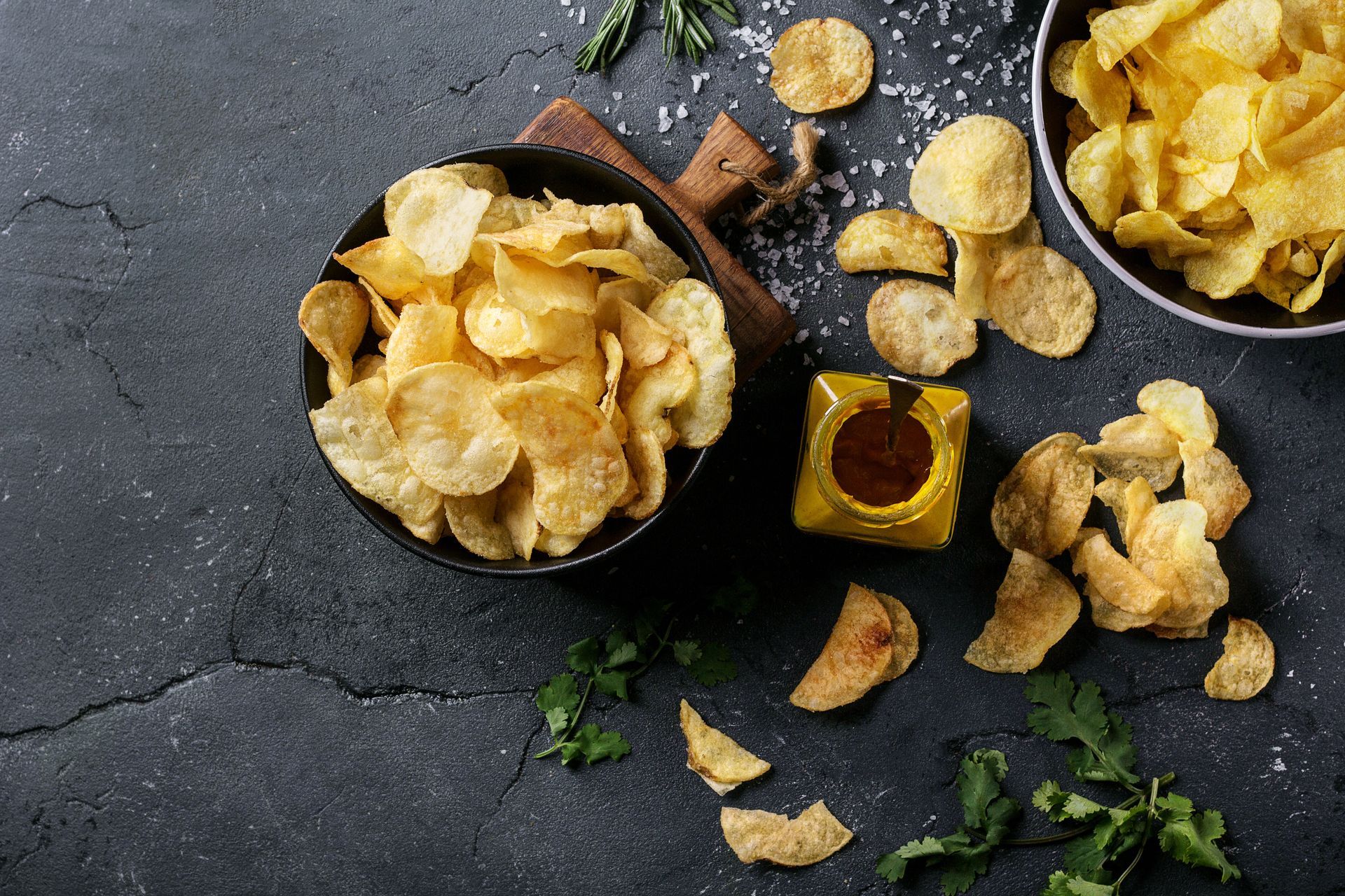 Bol de chips avec des morceaux éparpillés, petit pot de sauce et herbes aromatiques sur une surface en ardoise sombre
