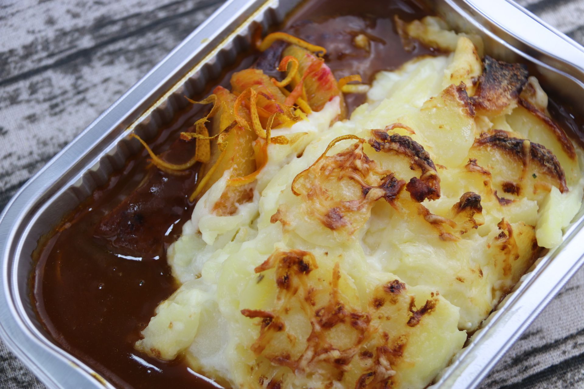 Gratin cuit au four dans un plat en aluminium, recouvert de purée de pommes de terre dorée et de carottes râpées, sur une table rustique
