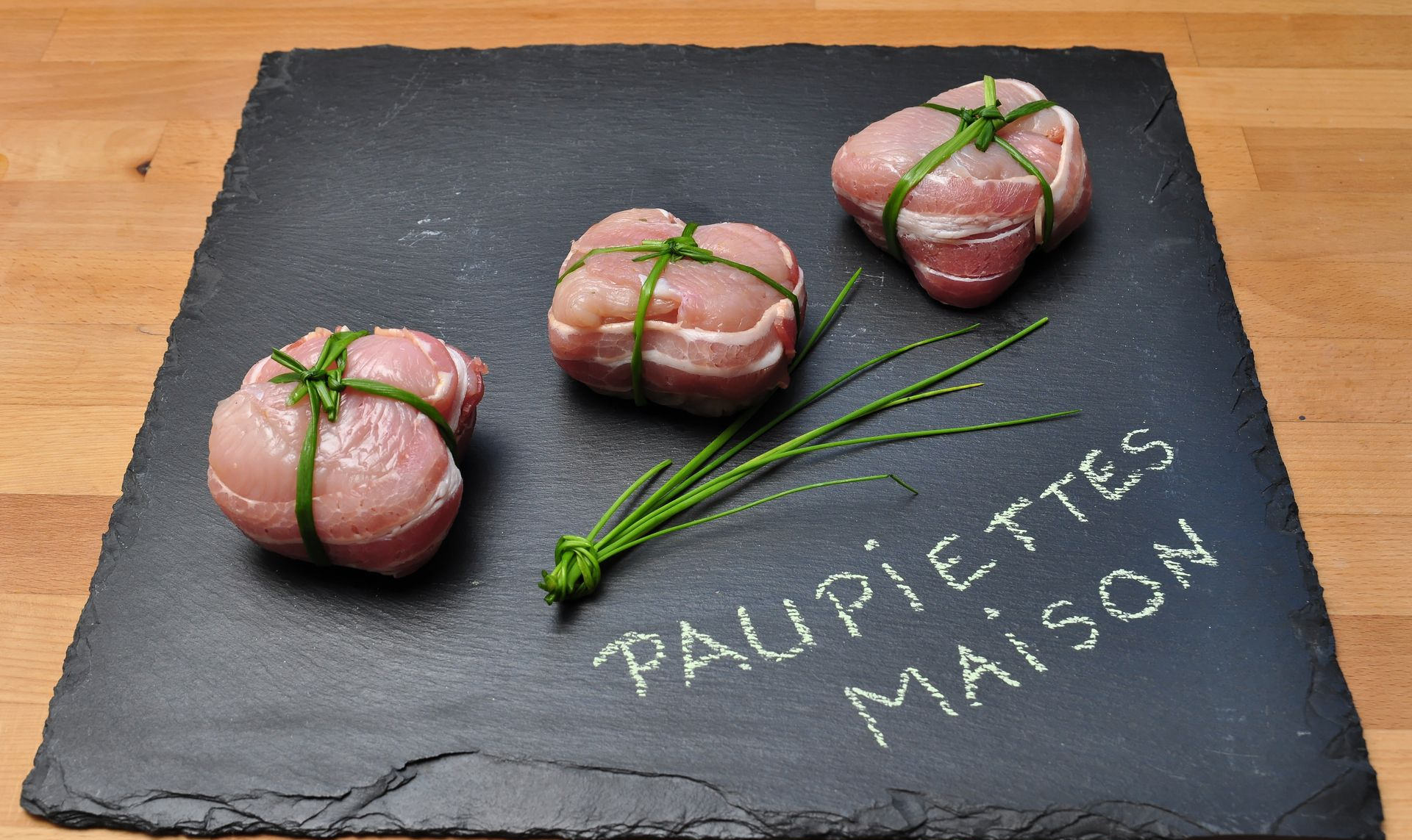 Trois paquets de viande crue ficelés sur une ardoise avec des herbes et l'inscription PAUPIETTES MAISON écrite à la farine