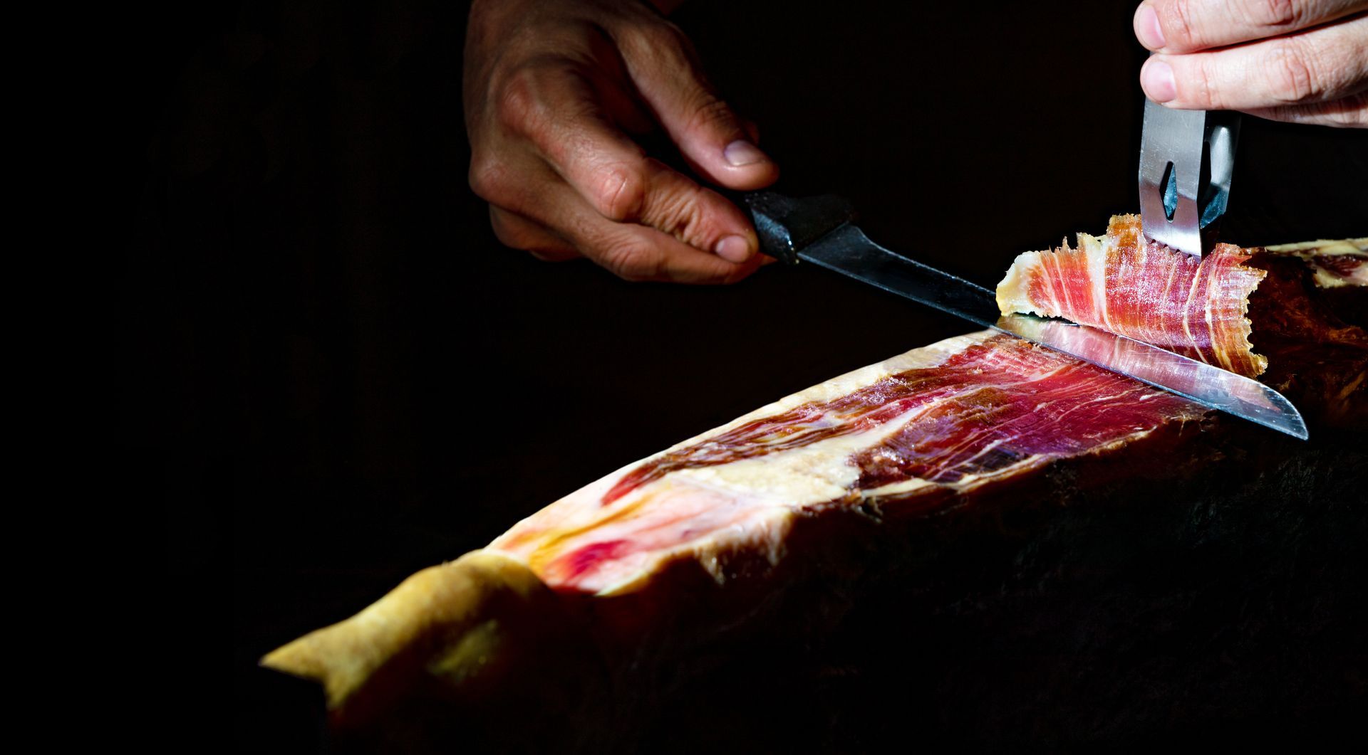 Des mains découpent un jambon cru au couteau sur un fond sombre.