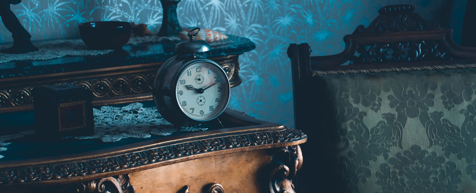 Une horloge ancienne ornée posée sur une table dans une pièce faiblement éclairée, avec une chaise à motifs en arrière-plan.