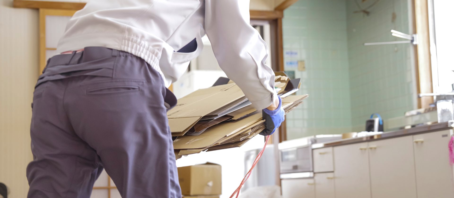 Une personne transporte une pile de cartons dans une pièce, probablement une cuisine. Elle porte des gants bleus et des vêtements de travail.