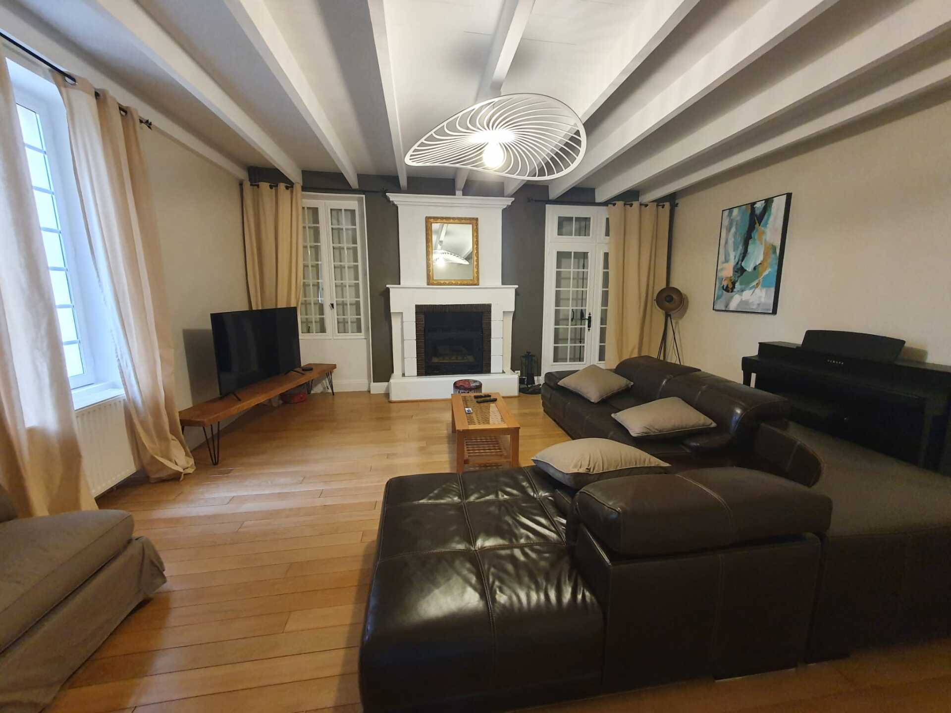 Plafond aux poutres apparentes blanches avec un lustre blanc accroché dessus, une cheminée au fond de la pièce et un grand canapé