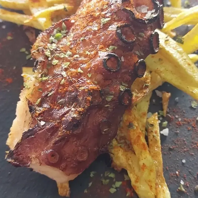 Pulpo a la parrilla sazonado con hierbas y pimentón, servido en un plato negro con finas tiras de patata fritas.