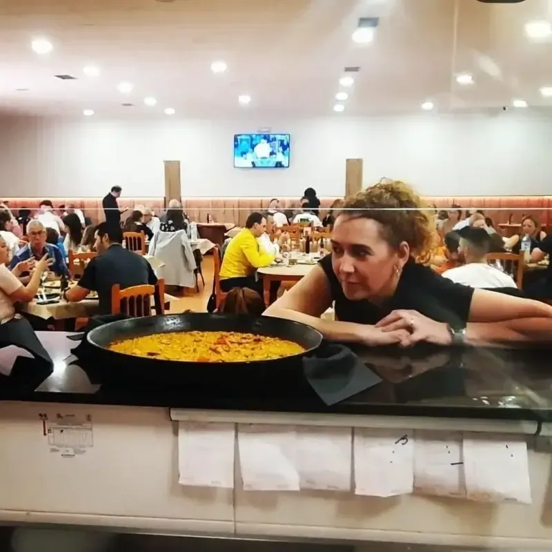 Una persona se apoya en la barra de un restaurante junto a una gran paella, con comensales al fondo.