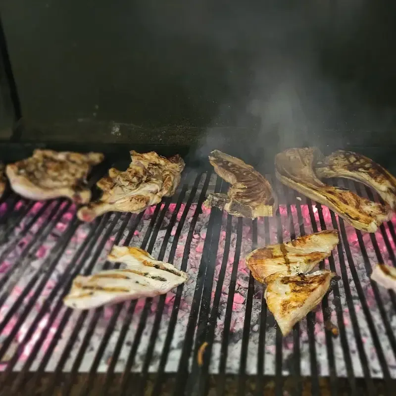 Diversos trozos de carne cocinándose en una parrilla de carbón caliente, con humo que se eleva.