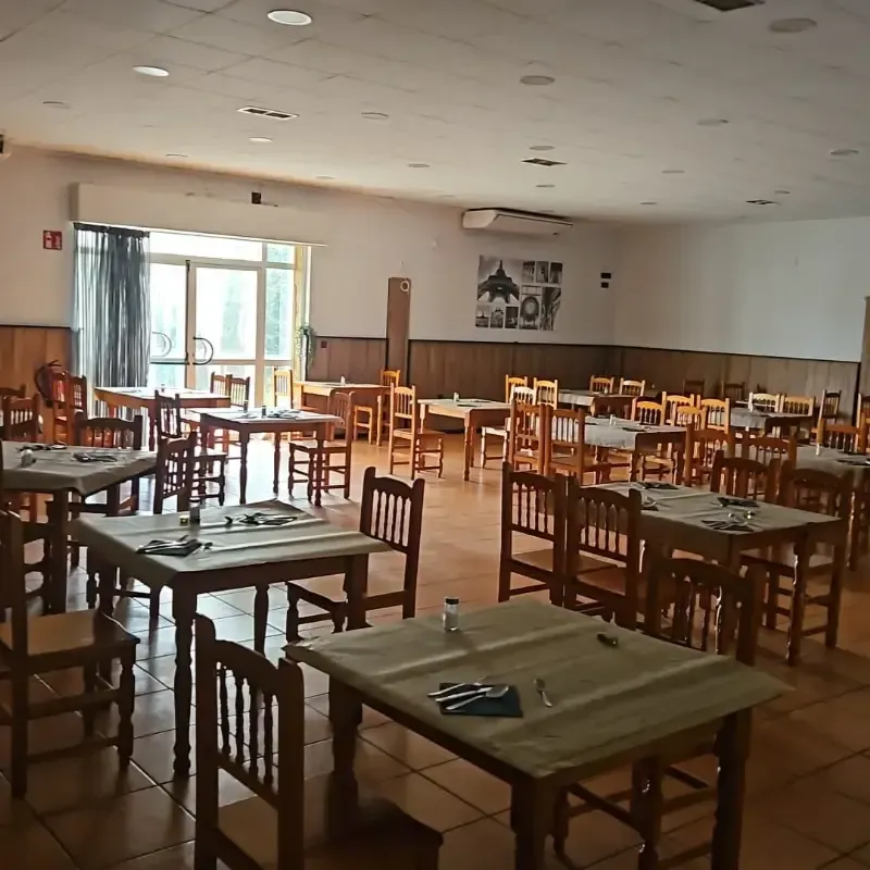 Un comedor amplio y vacío con mesas y sillas de madera, suelo de baldosas y un cuadro en la pared blanca del fondo.